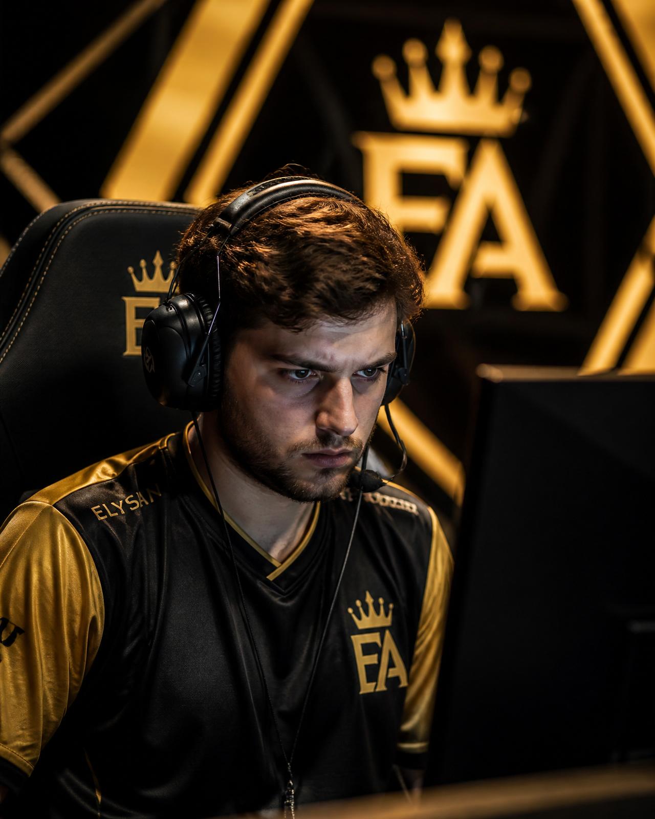 Male pro esports player in a black and gold jersey with a crown emblem, headset on, illuminated by monitor glow